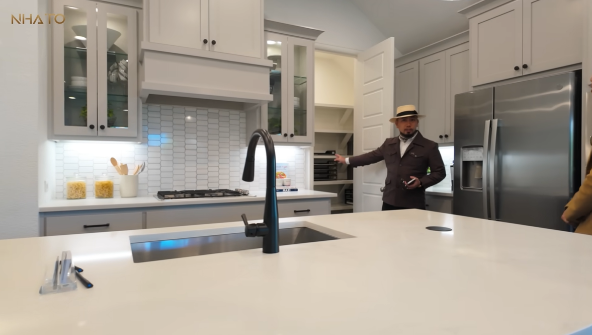 Nhà bếp hiện đại với tủ gỗ, bếp ga và vòi nước, người đàn ông đội mũ đứng chỉ vào phòng chứa đồ.