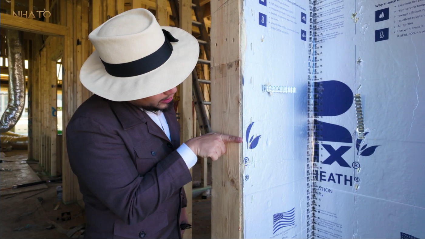 Người đàn ông mặc áo vest và đội mũ rộng vành đang chỉ vào tấm cách nhiệt trên công trường xây dựng.