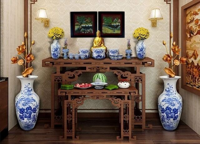 Bàn thờ gỗ chạm khắc với bình hoa sen, tượng Phật, đồ gốm sứ xanh và đèn tường vàng.