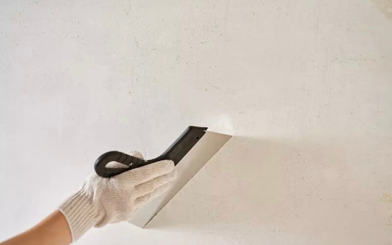 son-ba-tuong-la-gi Người đeo găng tay cầm dao xốp trát tường trắng, chuẩn bị bề mặt cho sơn hoặc sửa chữa.