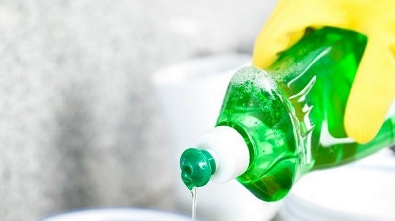 Chai nước rửa chén màu xanh lá cây đang được đổ ra, tay đeo găng cao su vàng.