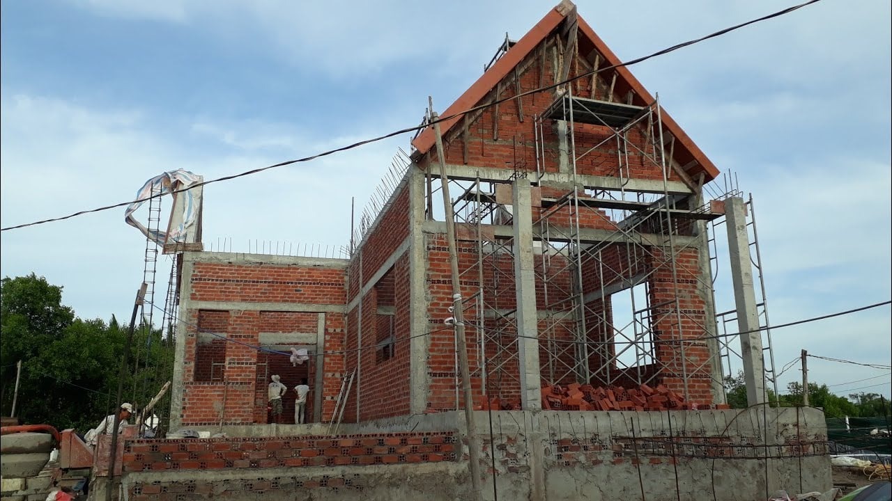 Nhà đang xây dựng với dàn giáo và tường gạch, trời xanh mây trắng.