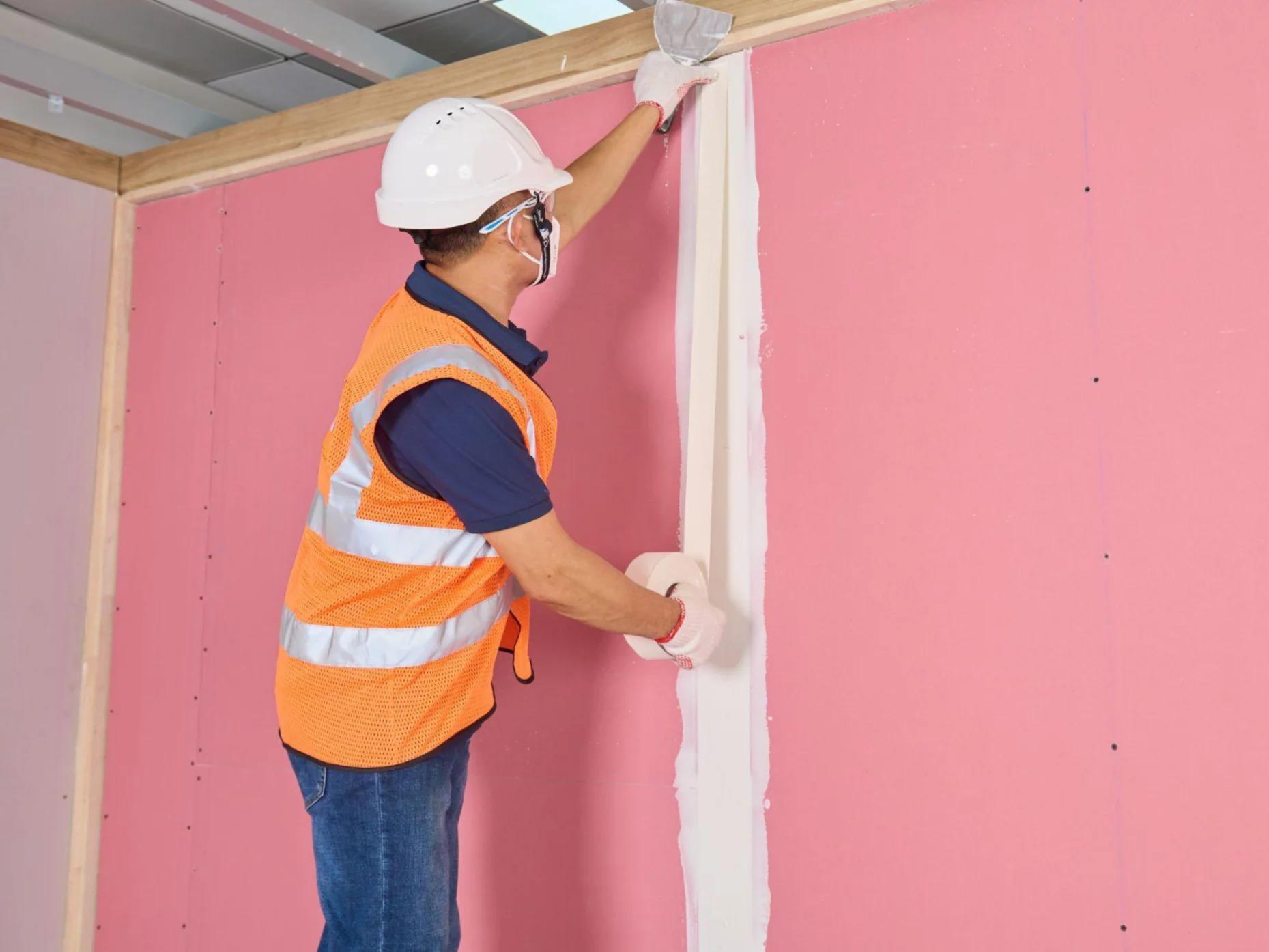 Người công nhân xây dựng đang dán băng trên tường thạch cao hồng, mặc áo bảo hộ và đội mũ bảo hiểm.