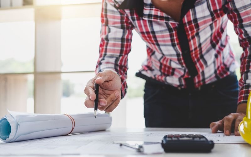 Người đàn ông kiểm tra bản vẽ kiến trúc trên bàn với bàn tay cầm bút, công cụ xây dựng và máy tính.