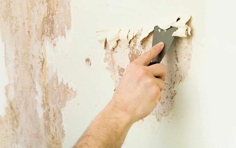 thi-cong-son-ba-buoc-2 Người đang cạo lớp sơn bong tróc trên tường bằng dao cạo kim loại.