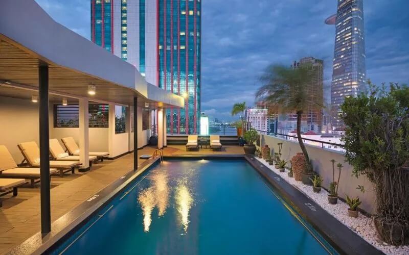 Hồ bơi trên sân thượng với ghế dài, tầm nhìn thành phố và tòa nhà cao tầng vào buổi chiều tối.