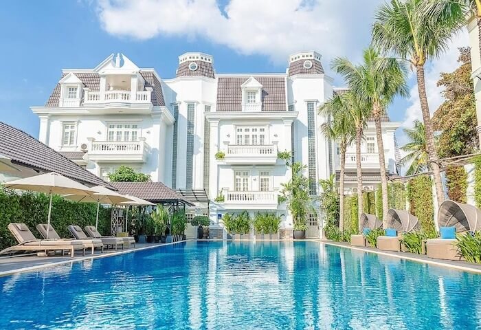 Biệt thự sang trọng với hồ bơi xanh, ghế nằm, cây cọ và trời trong xanh, tạo không gian nghỉ dưỡng đẳng cấp.