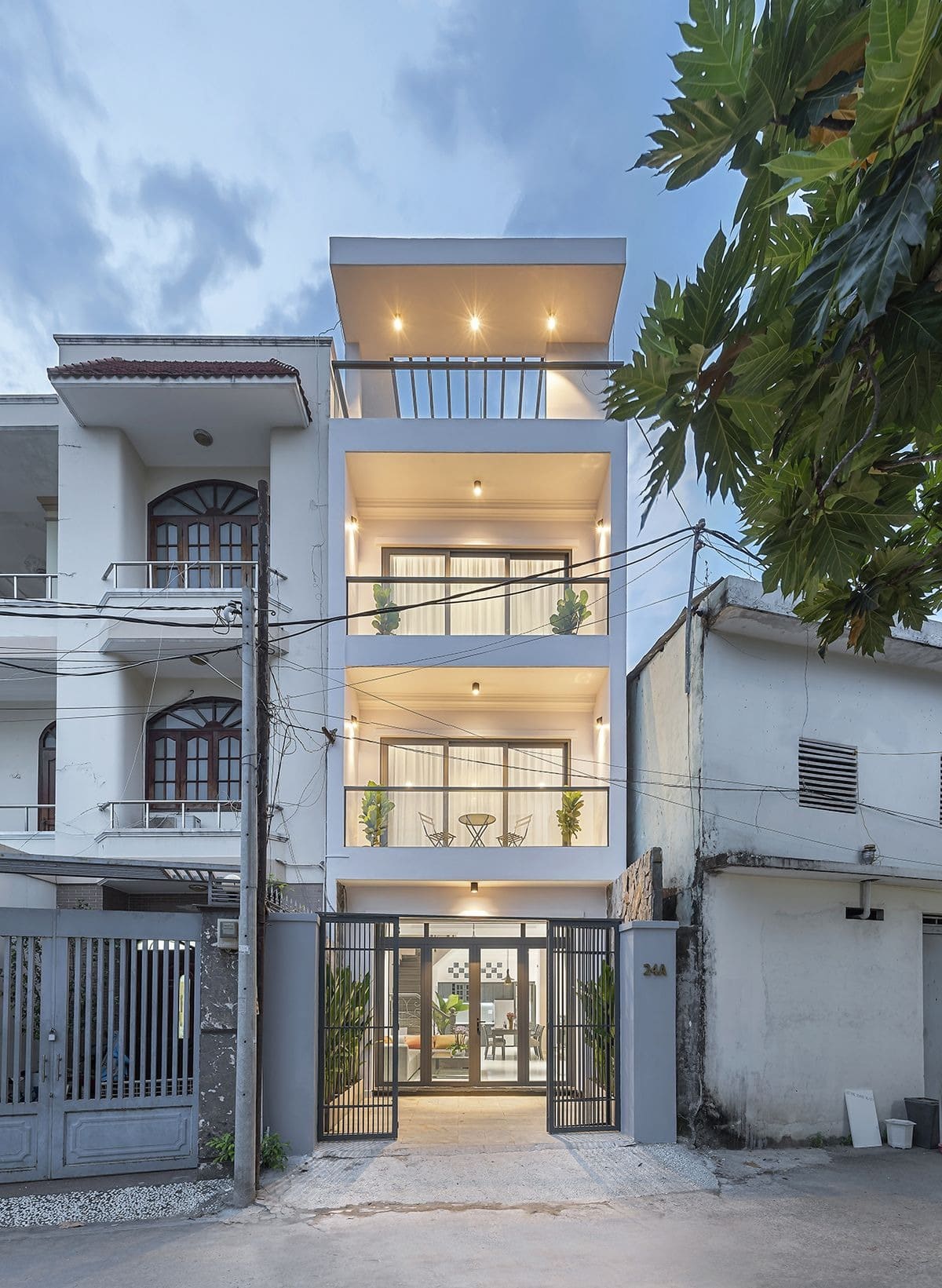 Nhà phố hiện đại 3 tầng với ban công, đèn chiếu sáng và cây xanh, nằm trên con đường yên tĩnh.