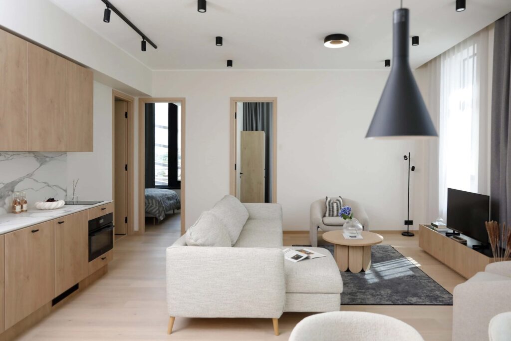 Phòng khách hiện đại với sofa xám, bàn gỗ, bếp mở, ánh sáng tự nhiên. Nội thất tối giản, tông màu trung tính.