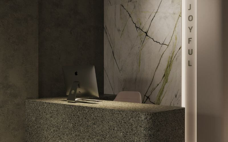 Quầy lễ tân hiện đại với máy tính iMac, tường đá cẩm thạch sang trọng, chữ Joyful bên cạnh.