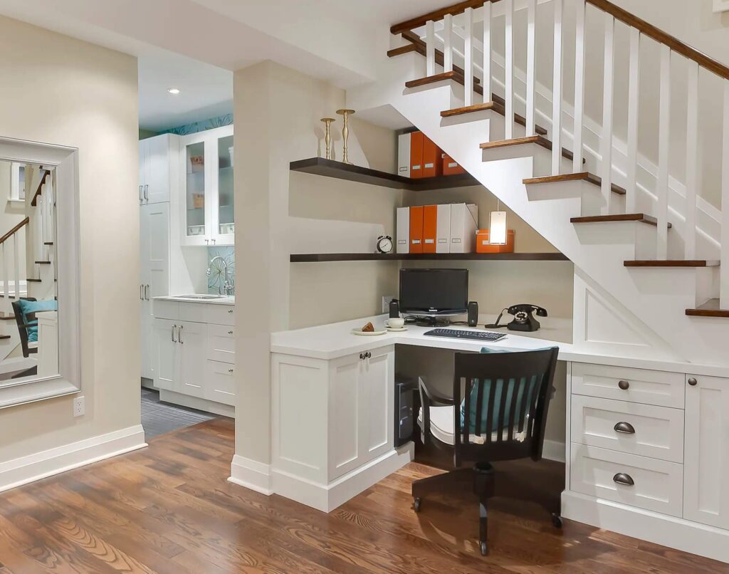 Góc làm việc nhỏ gọn dưới cầu thang với bàn trắng, máy tính, kệ sách và sàn gỗ.