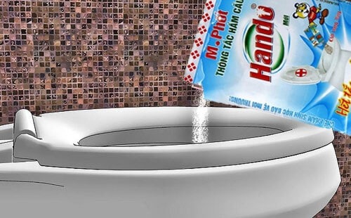 Gói bột thông cống Hando đang được đổ vào bồn cầu sứ trắng trên nền gạch nhà vệ sinh màu nâu.