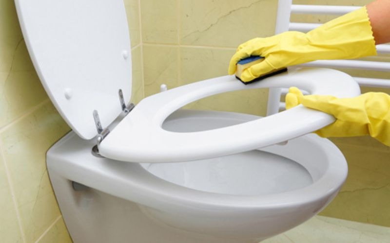 thuc-hien-ve-sinh-dung-cach-de-bao-ve-nap-nhua Người đeo găng tay cao su đang vệ sinh bồn cầu trong phòng tắm.
