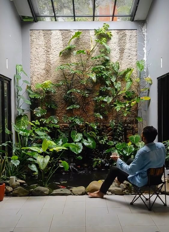 Người đàn ông thư giãn bên khu vườn nhỏ trong nhà với tường cây xanh và ao cá, tạo cảm giác bình yên.