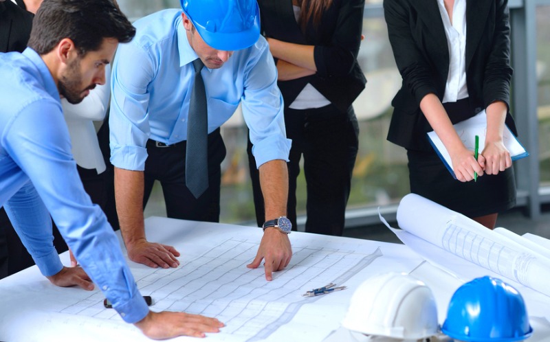 Nhóm kỹ sư và kiến trúc sư đang thảo luận kế hoạch xây dựng trên bàn có bản vẽ và mũ bảo hộ.