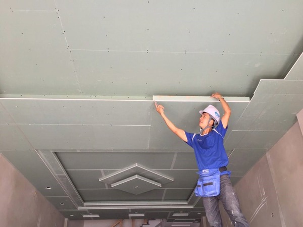 tran-thach-cao-la-gi Người thợ lắp đặt trần thạch cao trong nhà, mặc đồng phục xanh, đội mũ bảo hộ.