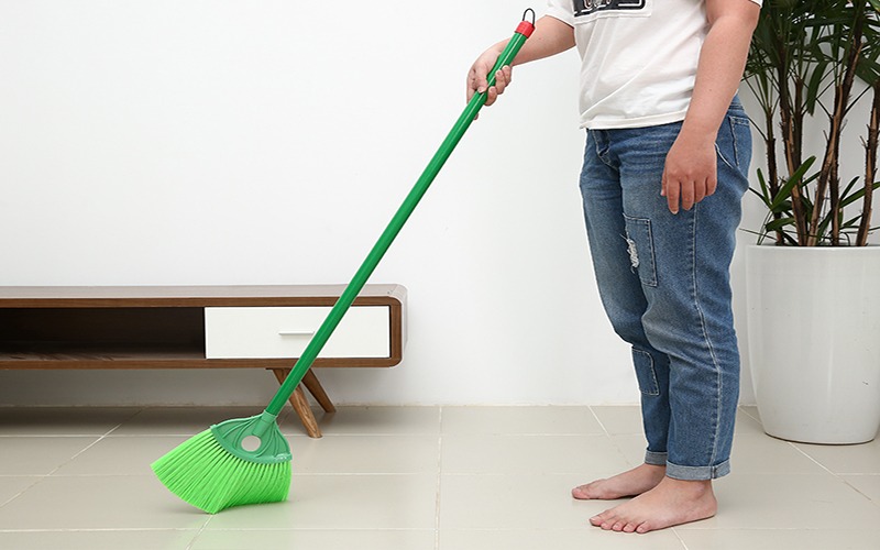 Người cầm chổi màu xanh lá quét sàn sáng sủa, cạnh kệ gỗ và cây xanh trong phòng sạch sẽ.