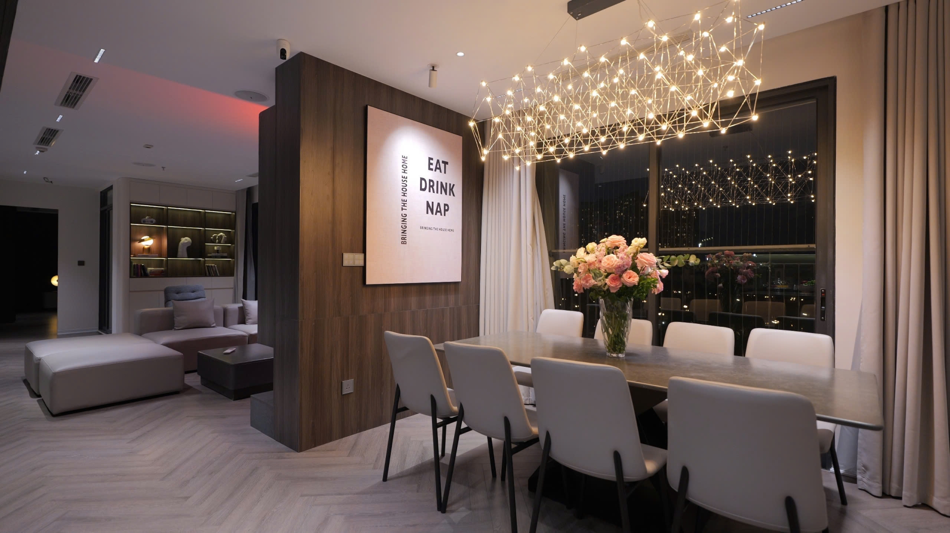 ub1wvnyi5gxjz0e2p6mr Phòng ăn hiện đại với đèn chùm sang trọng, bàn dài và bình hoa hồng trên bàn, không gian mở và tươi sáng.