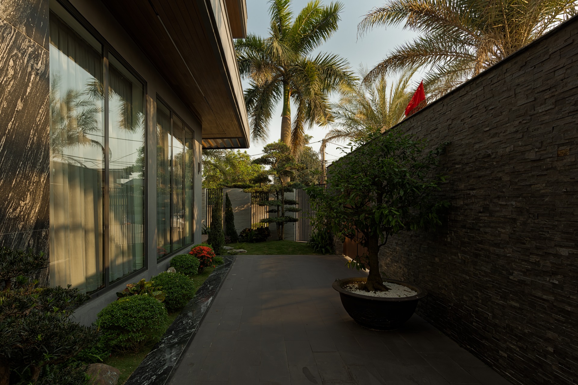 umi8686-enhanced-nr-edit Lối đi sân vườn với cây cối xanh tươi và nhà hiện đại, ánh nắng chiếu qua cửa kính lớn.