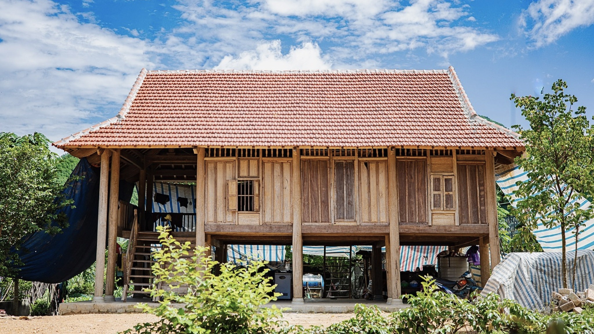 Nhà sàn truyền thống bằng gỗ với mái ngói đỏ, nằm giữa thiên nhiên xanh tươi, trời xanh mây trắng.