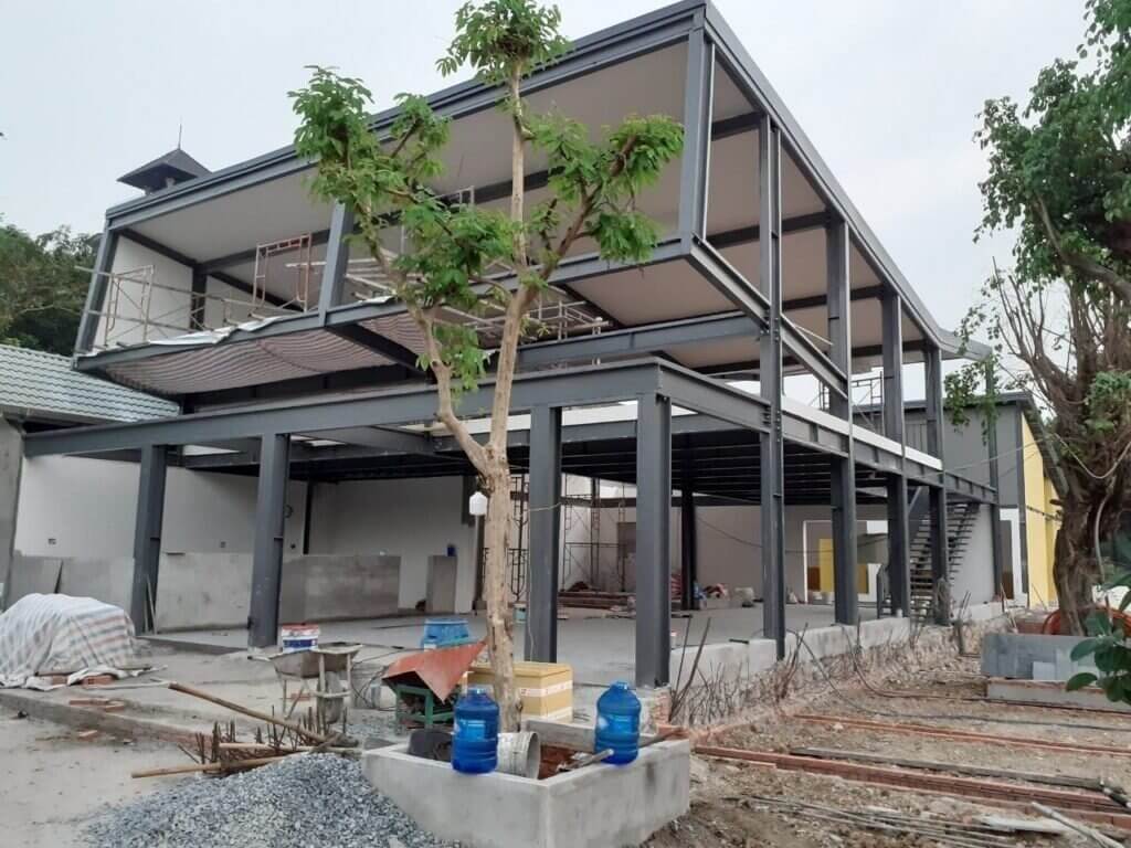 uu-diem-cua-nha-thep-tien-che-thoi-gian-thi-cong-nhanh-chong Khung nhà thép hai tầng đang được xây dựng với công trình và cây xanh xung quanh.