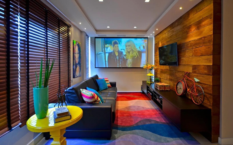 Phòng khách hiện đại với màn chiếu lớn, ghế sofa đen, tường gỗ và trang trí đa sắc màu.