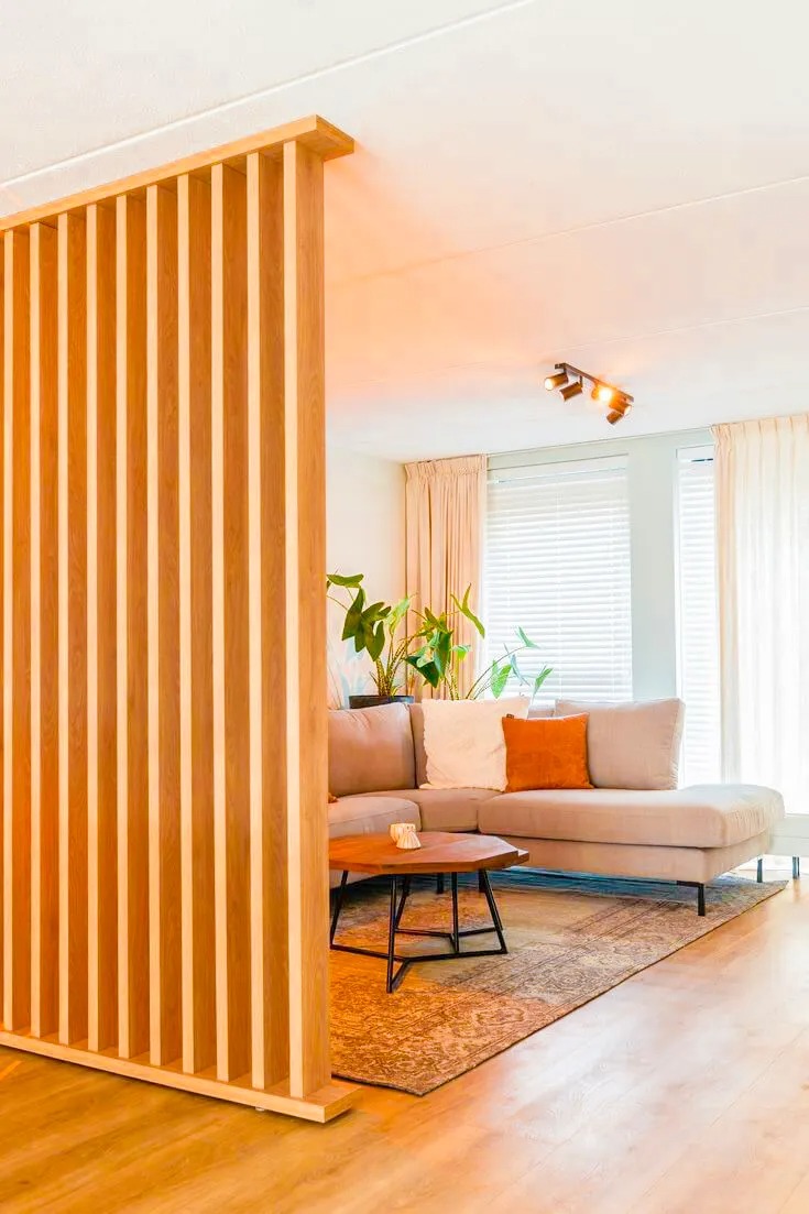 Phòng khách hiện đại với sofa góc, bàn gỗ, cây cảnh và vách ngăn gỗ tạo không gian ấm cúng.