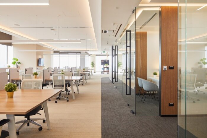 Văn phòng hiện đại với không gian mở, bàn làm việc gỗ, ghế xoay, cây xanh và vách kính ngăn cách phòng họp.