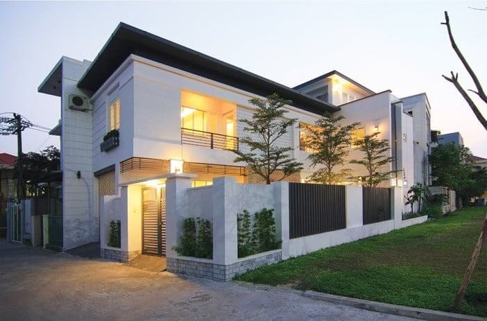 Ngôi nhà hiện đại với ánh đèn vàng, cây xanh và hàng rào cao vào buổi tối.