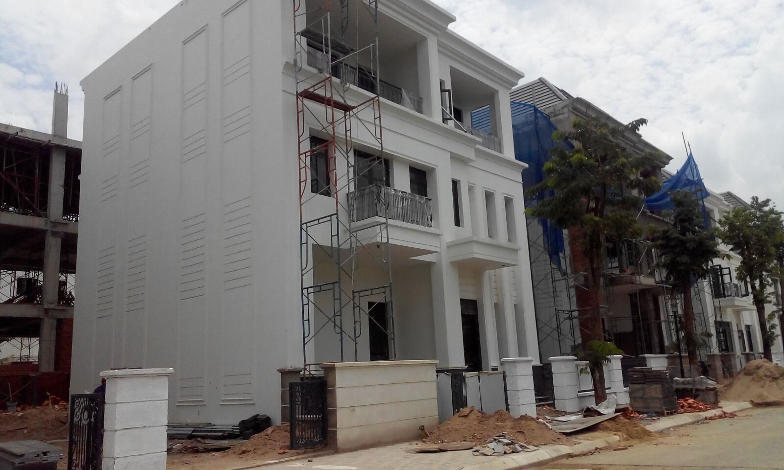 Nhà phố đang xây dựng với giàn giáo và hàng rào, cây xanh xung quanh, trời nhiều mây.