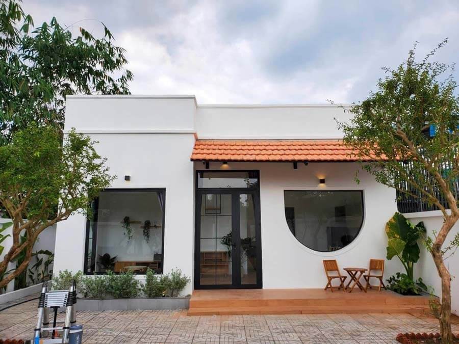 xac-dinh-nhu-cau-su-dung-va-phong-cach-thiet-ke Nhà hiện đại với mái ngói đỏ và cửa sổ lớn, bao quanh bởi cây xanh và bàn ghế ngoài trời, dưới bầu trời xanh.