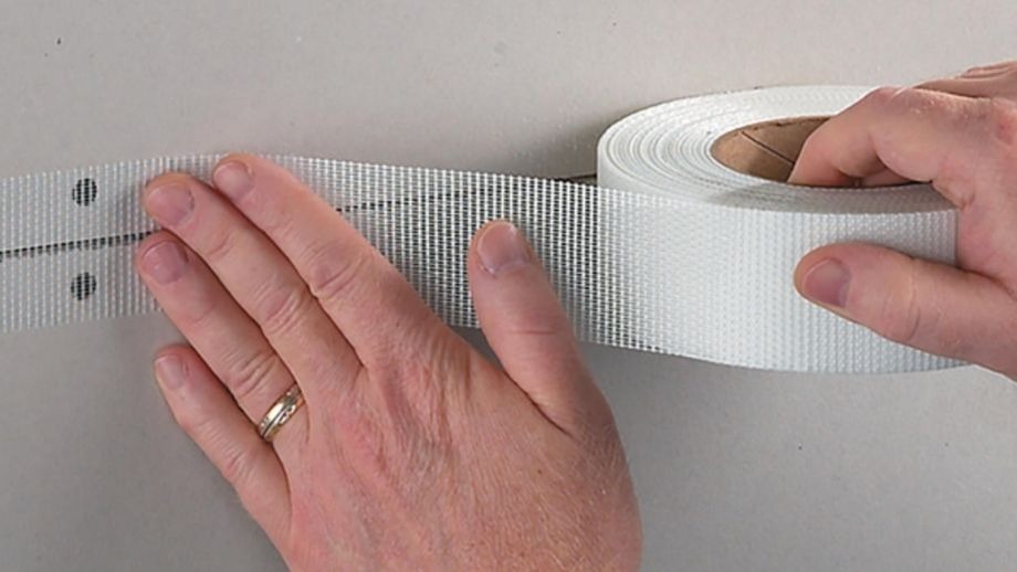 xu-ly-moi-noi-va-son-hoan-thien Cuộn băng keo lưới trắng được tay người cầm và căng ra, chuẩn bị sử dụng trên bề mặt tường.