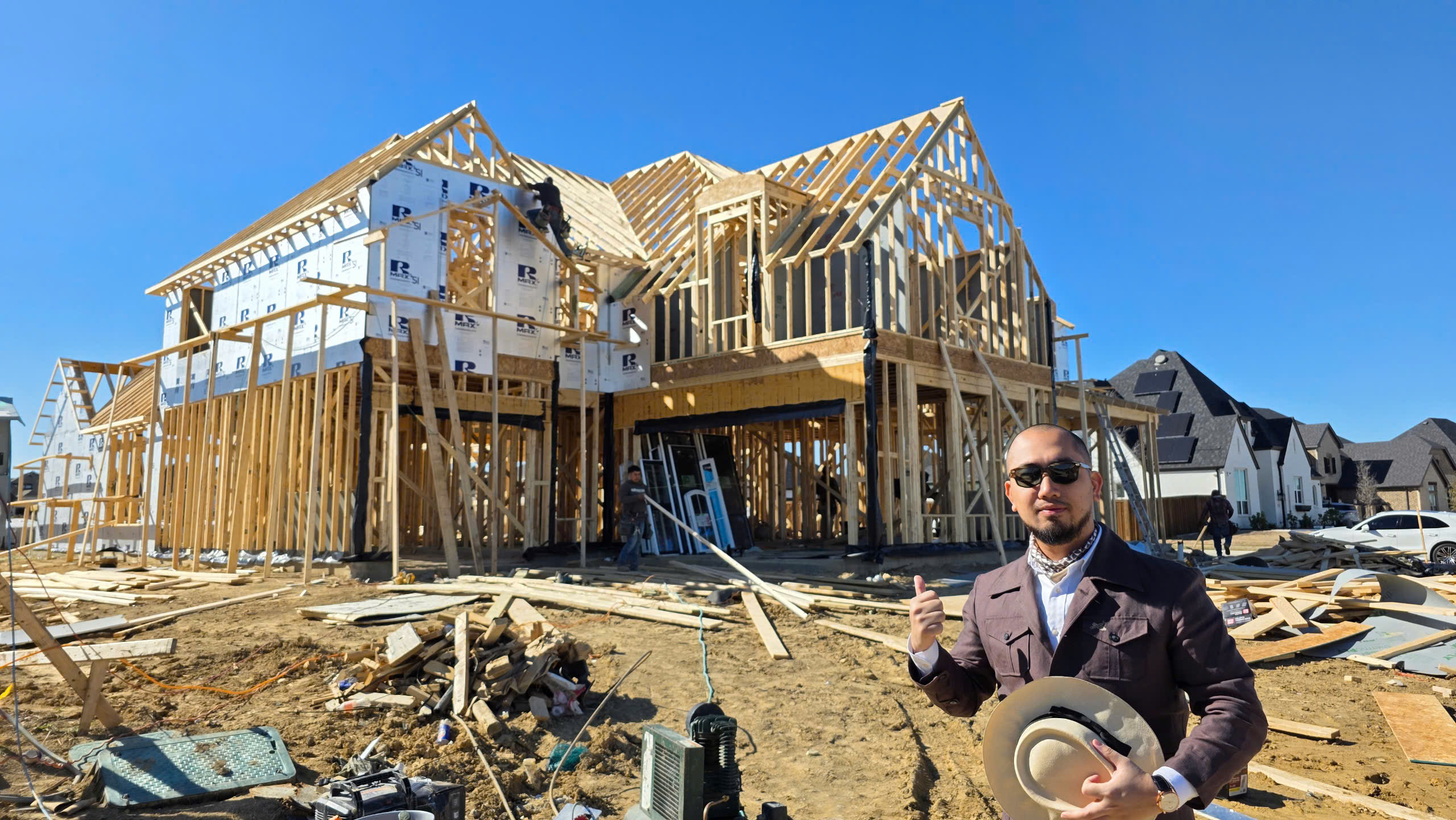 z6817939231705_b72614f408b61f19206c8d55537107d1 Người đàn ông đứng trước ngôi nhà đang xây dựng với kết cấu gỗ, trời trong xanh.