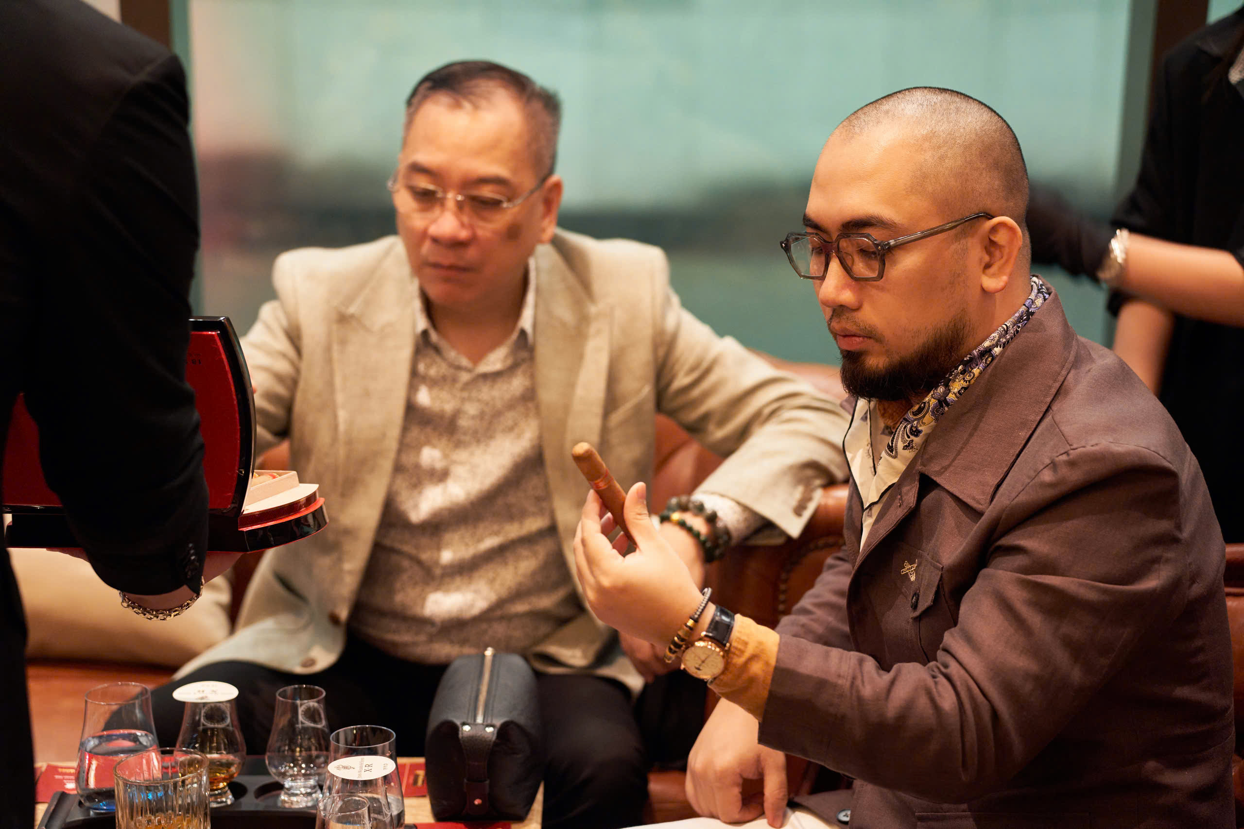 z6865896510912_955da34357c4e4f187a5d1897d3f63f1 Hai người đàn ông đang ngồi, một người cầm điếu xì gà và đồ uống trên bàn.