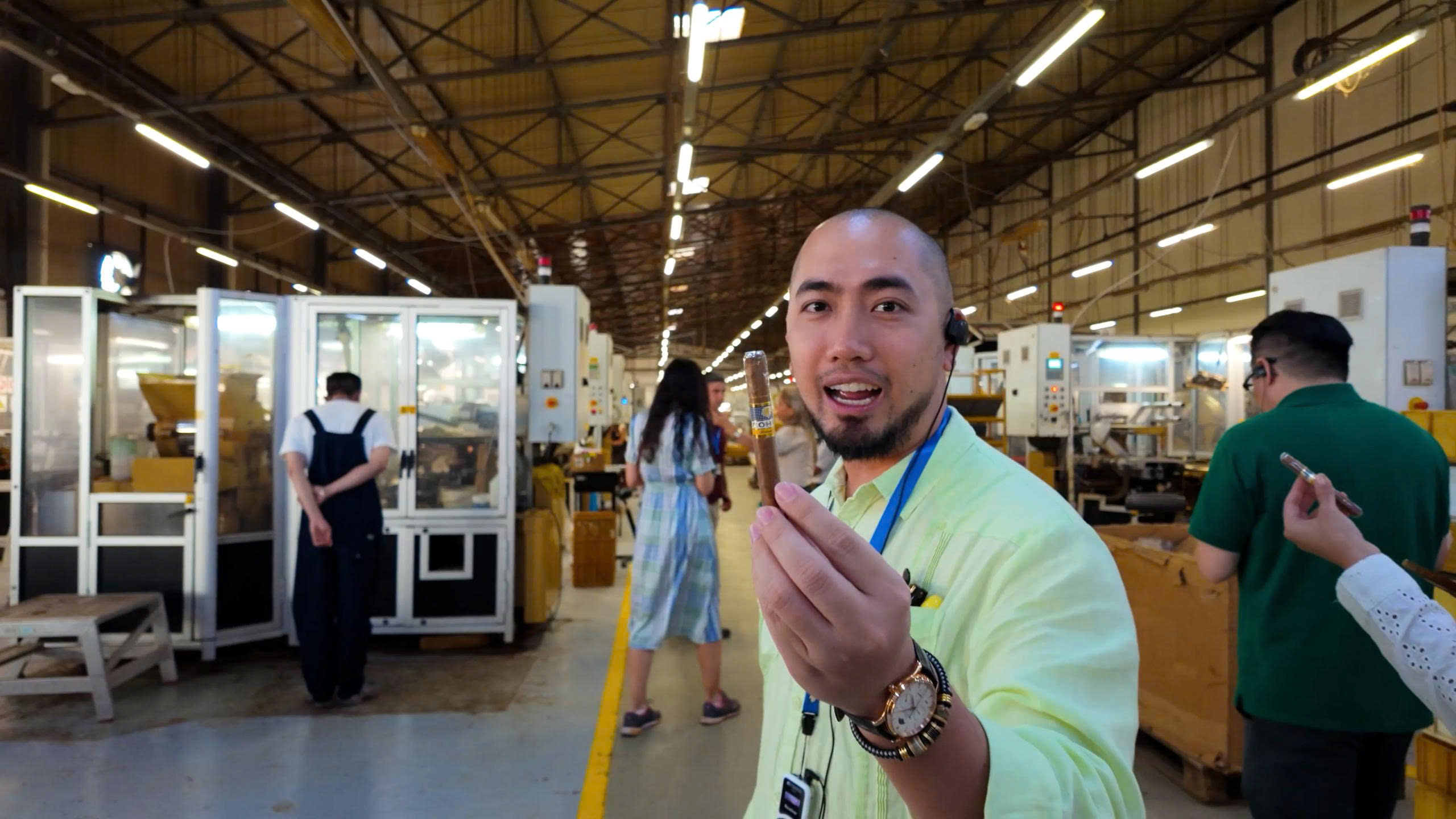 z6879709489971_9dac39032787f9e30d3a6fbc5946e173 Người đàn ông cầm điếu xì gà trong nhà máy sản xuất, nền là công nhân và máy móc.