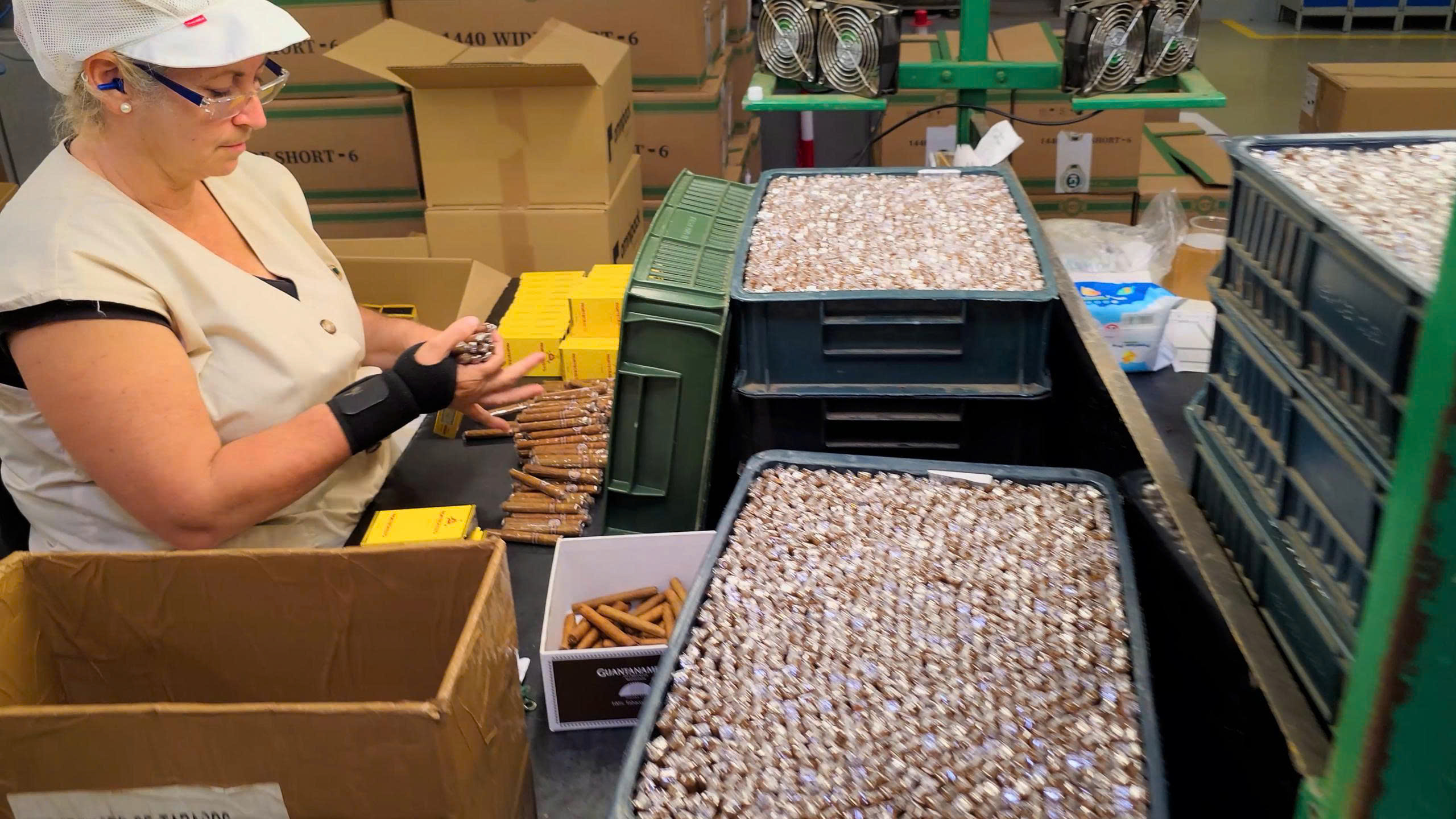 z6879709616145_de3cfce7070f4cc60514a9315ddb4b96 Người phụ nữ làm việc trong nhà máy sản xuất thuốc lá với nhiều hộp và thùng chứa xung quanh.