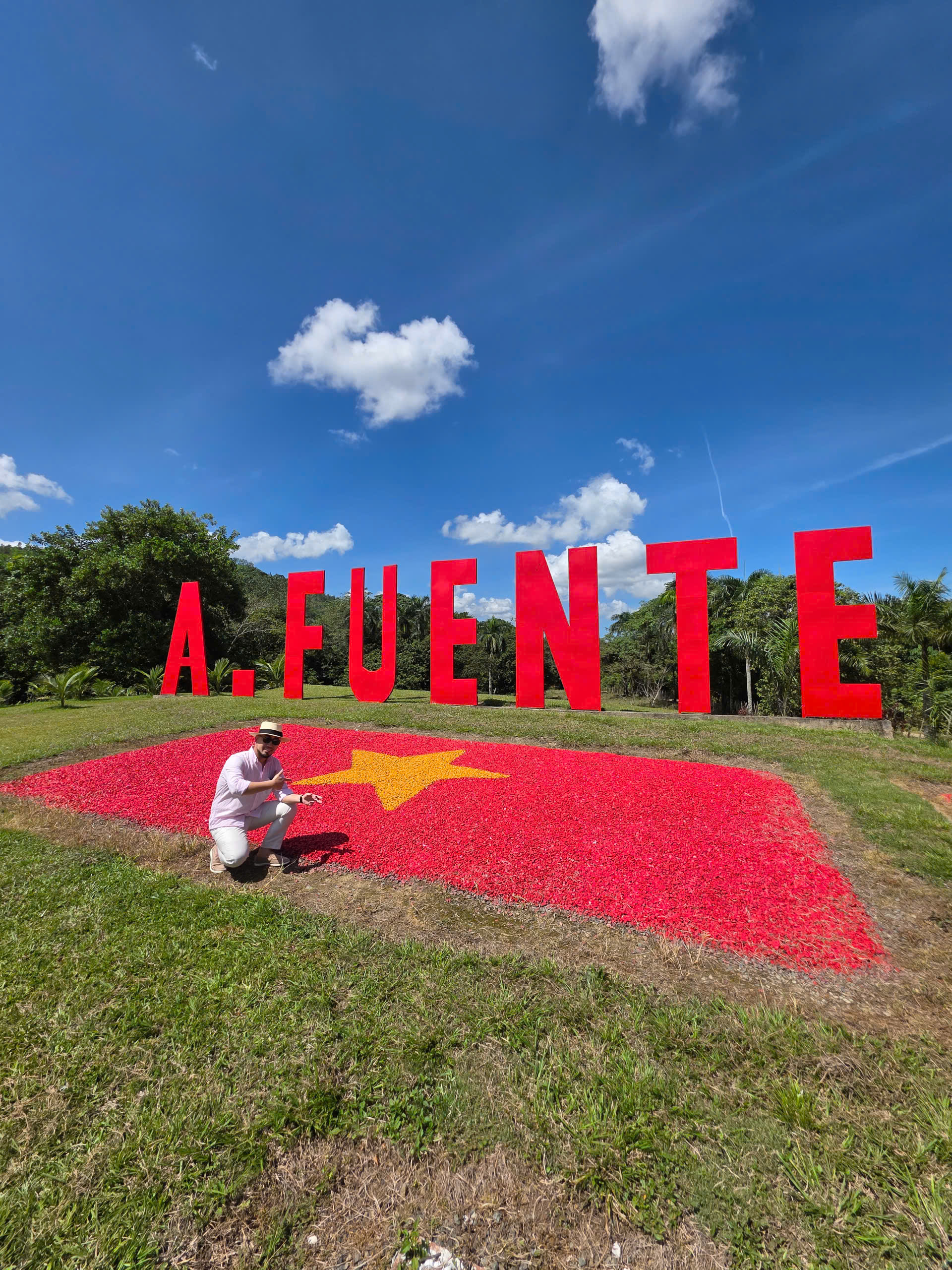 z6936179262096_f48eca389bb6a3e878f4175206686ade-1 Người đàn ông chụp ảnh trước khẩu hiệu A. FUENTE đỏ tươi, trời xanh trong, có cờ Việt Nam trên thảm hoa.